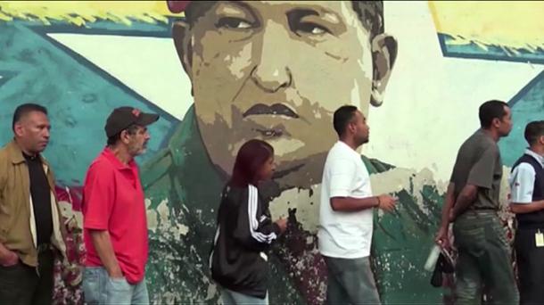 Varios venezolanos aguardan en fila delante una pintura de Nicolás Maduro.